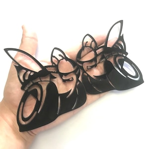 May include: Two black, laser-cut butterfly-shaped ornaments with intricate wing and body details. The ornaments are held in a hand against a white background. The cut-out design allows light to pass through, creating a delicate appearance.