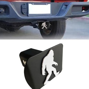 May include: Black trailer hitch cover featuring a white silhouette of Bigfoot. The hitch cover is rectangular with rounded edges and is designed to fit standard trailer hitches. The image shows the hitch cover installed on a red truck.