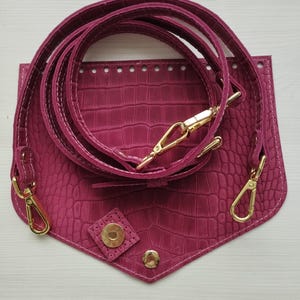 Pode incluir: Uma aba de bolsa magenta com textura de pele de crocodilo, apresentando uma fileira de furos pré-perfurados ao longo da borda superior, um fecho magnético dourado e uma alça de ombro ajustável combinando com mosquetões giratórios dourados.