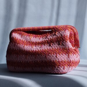 Puede incluir: Un bolso de mano rectangular de ganchillo con un vibrante patr&oacute;n de rayas horizontales en tonos rosa, coral y naranja. El bolso presenta una abertura de marco estructurado con un asa a juego y una construcci&oacute;n de hilo tejido texturizado.