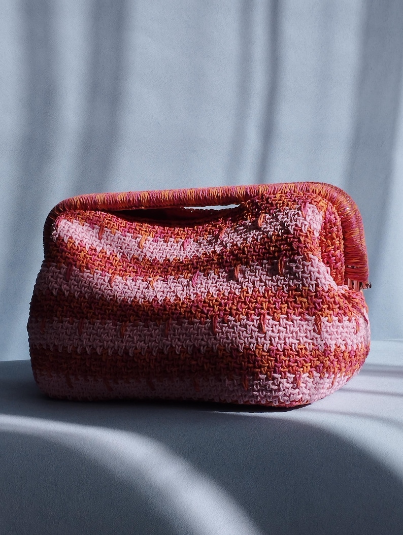 Puede incluir: Un bolso de mano rectangular de ganchillo con un vibrante patr&oacute;n de rayas horizontales en tonos rosa, coral y naranja. El bolso presenta una abertura de marco estructurado con un asa a juego y una construcci&oacute;n de hilo tejido texturizado.