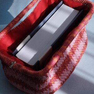 Puede incluir: Un bolso rectangular de ganchillo con un patr&oacute;n de rayas en tonos rosa y naranja. El bolso tiene una abertura de marco estructurado con un cierre de metal y est&aacute; forrado con una tela roja s&oacute;lida. Un libro grueso de tapa dura est&aacute; dentro.