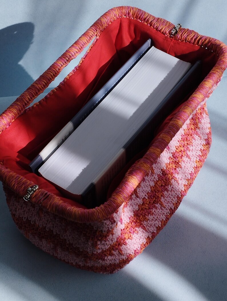 Puede incluir: Un bolso rectangular de ganchillo con un patr&oacute;n de rayas en tonos rosa y naranja. El bolso tiene una abertura de marco estructurado con un cierre de metal y est&aacute; forrado con una tela roja s&oacute;lida. Un libro grueso de tapa dura est&aacute; dentro.