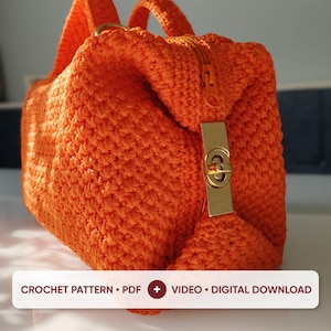 May include: A bright orange crochet handbag featuring a textured stitch pattern, two top handles, and a gold-tone metal twist-lock closure. The image includes a white rectangular label with the text: CROCHET PATTERN • PDF + VIDEO • DIGITAL DOWNLOAD.