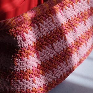Puede incluir: Un bolso tote rectangular tejido con un patr&oacute;n de ganchillo texturizado en rayas diagonales alternas de rosa claro, rosa brillante y naranja. El bolso tiene un borde superior reforzado en un tejido a juego y un forro interior de tela roja s&oacute;lida.