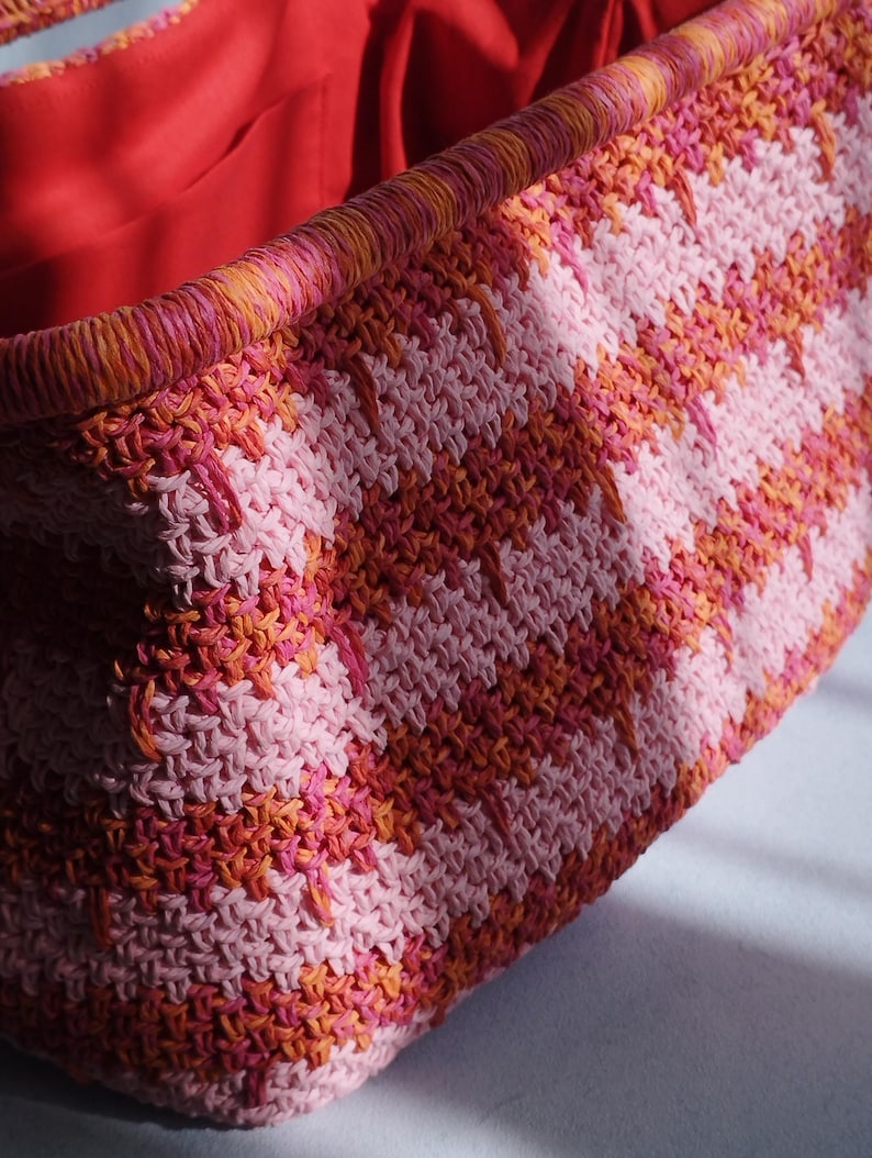 Puede incluir: Un bolso tote rectangular tejido con un patr&oacute;n de ganchillo texturizado en rayas diagonales alternas de rosa claro, rosa brillante y naranja. El bolso tiene un borde superior reforzado en un tejido a juego y un forro interior de tela roja s&oacute;lida.