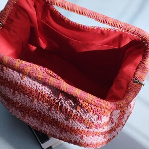 Puede incluir: Un bolso de mano rectangular de ganchillo con un patr&oacute;n de rayas vibrantes en tonos rosa, naranja y coral. El bolso tiene una abertura de marco estructurado y est&aacute; forrado con una tela roja s&oacute;lida, con un bolsillo abierto en el lateral.