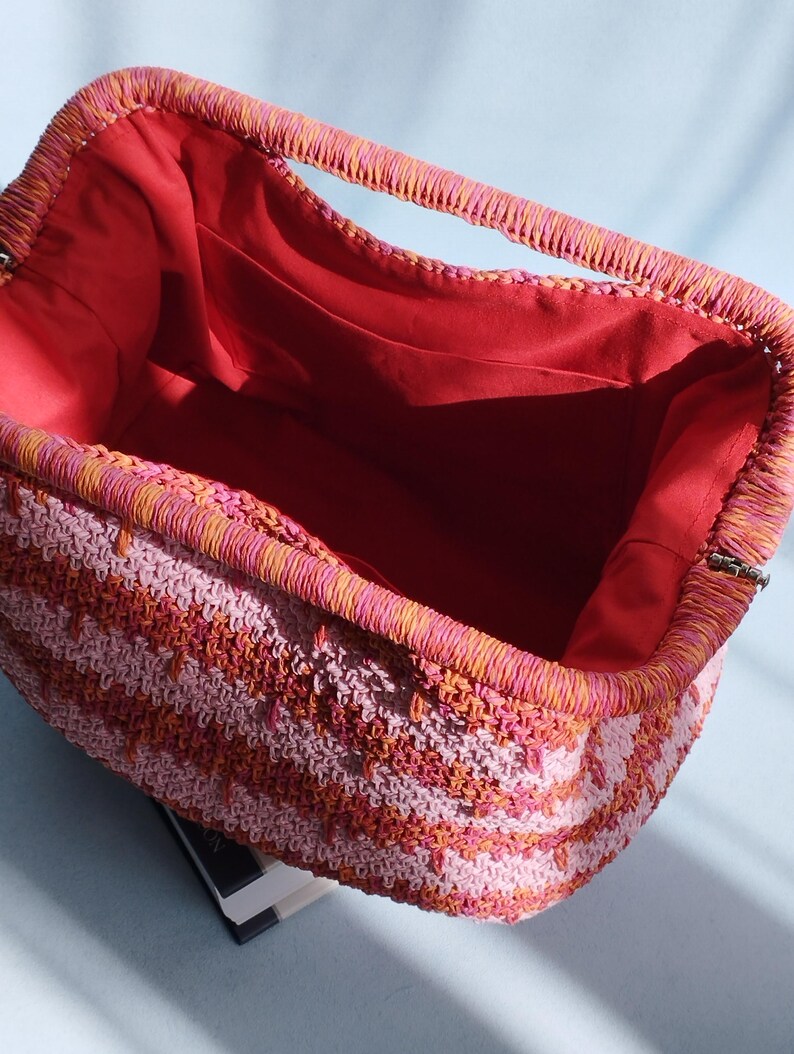 Puede incluir: Un bolso de mano rectangular de ganchillo con un patr&oacute;n de rayas vibrantes en tonos rosa, naranja y coral. El bolso tiene una abertura de marco estructurado y est&aacute; forrado con una tela roja s&oacute;lida, con un bolsillo abierto en el lateral.