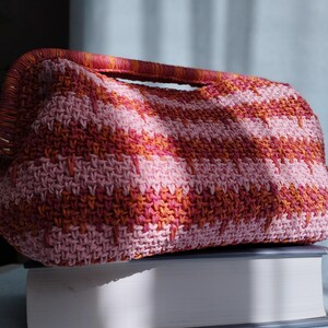 Puede incluir: Un bolso de mano rectangular de ganchillo con un patr&oacute;n de rayas horizontales en tonos rosa claro, rosa oscuro y naranja. El bolso tiene un asa superior estructurada integrada en el dise&ntilde;o, con una puntada texturizada visible en el exterior.