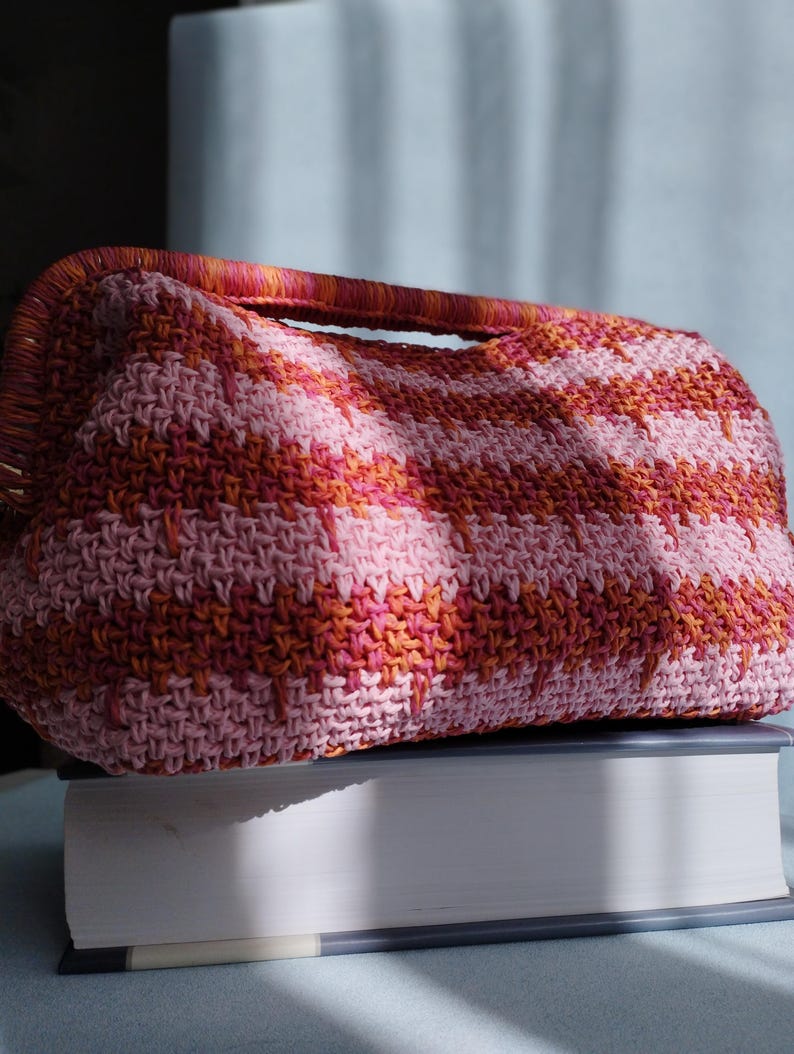 Puede incluir: Un bolso de mano rectangular de ganchillo con un patr&oacute;n de rayas horizontales en tonos rosa claro, rosa oscuro y naranja. El bolso tiene un asa superior estructurada integrada en el dise&ntilde;o, con una puntada texturizada visible en el exterior.