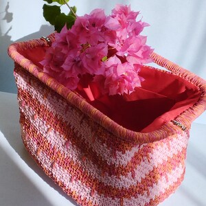 Puede incluir: Una cesta rectangular de ganchillo con un patr&oacute;n de chevr&oacute;n en tonos rosa y naranja. La cesta tiene un borde estructurado y un forro de tela roja lisa. Un ramo de flores de buganvilla de color rosa brillante se encuentra en su interior.