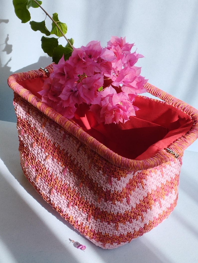 Puede incluir: Una cesta rectangular de ganchillo con un patr&oacute;n de chevr&oacute;n en tonos rosa y naranja. La cesta tiene un borde estructurado y un forro de tela roja lisa. Un ramo de flores de buganvilla de color rosa brillante se encuentra en su interior.