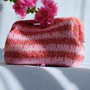 Puede incluir: Un bolso de mano rectangular de ganchillo con un patr&oacute;n de rayas horizontales en tonos rosa claro, coral y naranja. El bolso tiene una abertura de marco estructurado en la parte superior y est&aacute; decorado con un racimo de flores de buganvilla rosa.