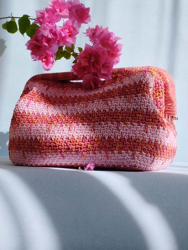 Puede incluir: Un bolso de mano rectangular de ganchillo con un patr&oacute;n de rayas horizontales en tonos rosa claro, coral y naranja. El bolso tiene una abertura de marco estructurado en la parte superior y est&aacute; decorado con un racimo de flores de buganvilla rosa.