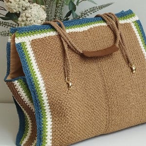 Könnte beinhalten: Eine große, handgefertigte Häkeltasche in Naturbeige. Die Tasche hat einen blau, weiß und grün gestreiften Rand. Sie hat braune Seilgriffe mit einem braunen Ledergriff und goldfarbener Hardware.