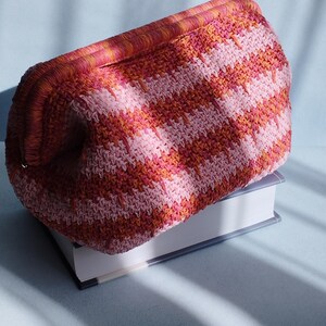 Puede incluir: Un bolso de mano de ganchillo con un patr&oacute;n de chevr&oacute;n texturizado y multicolor en tonos rosa, coral y naranja. El bolso tiene una estructura rectangular con un borde superior tejido a juego y un cuerpo de forma redondeada.