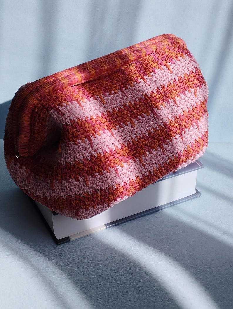 Puede incluir: Un bolso de mano de ganchillo con un patr&oacute;n de chevr&oacute;n texturizado y multicolor en tonos rosa, coral y naranja. El bolso tiene una estructura rectangular con un borde superior tejido a juego y un cuerpo de forma redondeada.