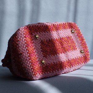Puede incluir: Un bolso de mano rectangular de ganchillo con un vibrante patr&oacute;n de bloques de color en tonos rosa, naranja y rosa claro. La base del bolso est&aacute; equipada con cuatro pies de metal dorado pulido, lo que proporciona estabilidad y estructura.