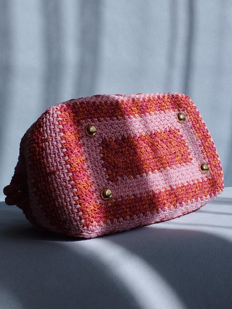 Puede incluir: Un bolso de mano rectangular de ganchillo con un vibrante patr&oacute;n de bloques de color en tonos rosa, naranja y rosa claro. La base del bolso est&aacute; equipada con cuatro pies de metal dorado pulido, lo que proporciona estabilidad y estructura.