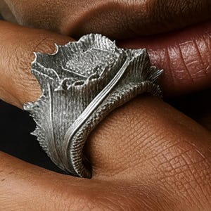 May include: A close-up of a silver ring with a textured, organic design. The ring appears to be made of metal and features a detailed, leaf-like shape. The ring is worn on a finger, with the skin visible in the frame.