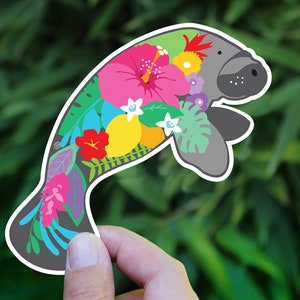 A cute, gray manatee sticker with a floral design. The manatee is outlined in white and has a colorful floral pattern of pink, yellow, red, blue, green, and white flowers and leaves.
