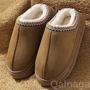 Puede incluir: Un par de zapatillas marrones de ante con una suela gruesa de color marrón claro. El interior está forrado con piel sintética blanca. El borde superior presenta una banda decorativa con un patrón geométrico en amarillo, naranja y blanco. Estas zapatillas son para uso en interiores.