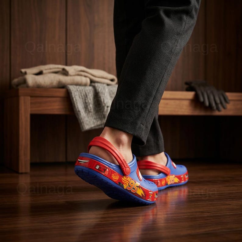 May include: Blue and red clogs featuring a Lightning McQueen design. The clogs have a red strap and the number 95 on the side. The shoes are worn on a wooden floor.
