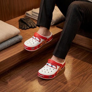 Puede incluir: Zuecos Crocs blancos y rojos de Lightning McQueen con un dise&ntilde;o de coche de dibujos animados. Los zuecos tienen una correa roja, detalles rojos y el n&uacute;mero 95 en el lateral. La persona lleva pantalones negros.