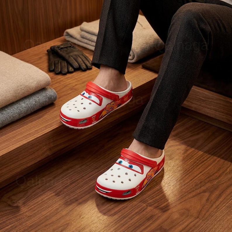 Puede incluir: Zuecos Crocs blancos y rojos de Lightning McQueen con un dise&ntilde;o de coche de dibujos animados. Los zuecos tienen una correa roja, detalles rojos y el n&uacute;mero 95 en el lateral. La persona lleva pantalones negros.