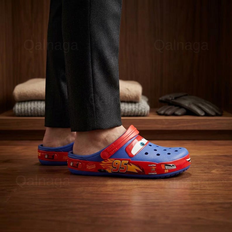 May include: Blue and red Lightning McQueen Crocs clogs. The clogs feature a red strap and a design of the character Lightning McQueen from the movie Cars. The number 95 is visible on the side of the shoe. The clogs are made of a lightweight material.