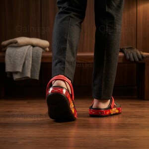May include: Red and black sandals with a decorative design. The sandals have a red strap and a black base with a colorful pattern. The person is wearing dark gray pants and walking on a wooden floor.