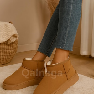 Op de afbeelding: Bruine enkellaarsjes met een dikke zool en een zacht, donzig interieur. De laarzen hebben het woord "Qalnaga" op de zijkant gedrukt. De laarzen worden gedragen met een blauwe jeans.