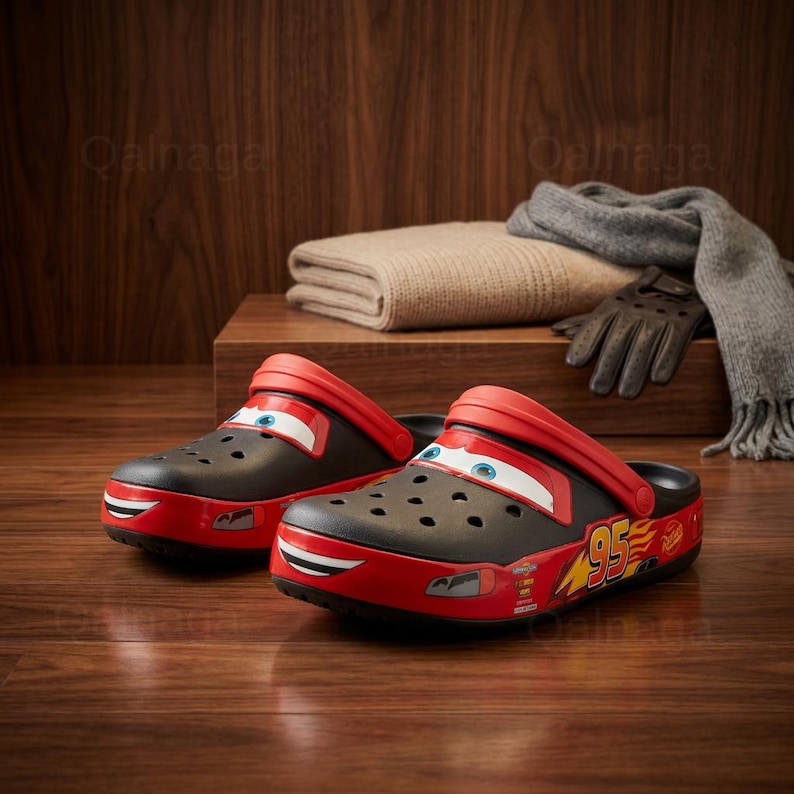 May include: Black and red Lightning McQueen Crocs clogs with a cartoon face design. The clogs have a red strap and a red bumper with the number 95 and other graphics. The shoes are on a wooden surface.