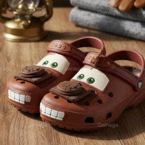 Könnte beinhalten: Ein Paar braune Clogs mit einem Cartoon-Auto-Design. Die Clogs haben eine weiße Front mit grünen Augen, einen braunen Oberteil und ein weißes, zahniges Grinsen. Die Schuhe sind aus einem gummiartigen Material gefertigt und auf Komfort ausgelegt.