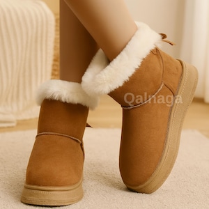 Puede incluir: Botines marrones con ribete de piel sintética color crema. Las botas tienen una puntera redondeada y una suela gruesa de color marrón claro. La palabra "Qalaga" está impresa en el lateral de la bota.