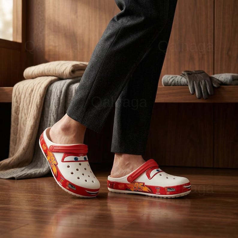 Puede incluir: Zuecos Crocs blancos y rojos de Lightning McQueen. Los zuecos presentan una correa roja, el n&uacute;mero 95 y un dise&ntilde;o de cara de dibujos animados. Los zapatos tienen m&uacute;ltiples orificios de ventilaci&oacute;n y los lleva una persona con pantalones grises.