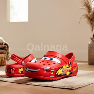 以下が含まれることがあります： 笑顔の顔のデザイン、青い目、側面に95の数字が付いた、赤いライトニング・マックイーンをテーマにしたクロッグ。クロッグにはストラップが付いており、ゴムのような素材でできています。靴はベージュのラグの上にあります。