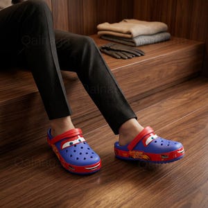 May include: Blue and red cartoon-themed clogs. The clogs feature a red strap, a cartoon car face on the front, and the number 95 on the side. The shoes are worn by a person sitting on a wooden surface.