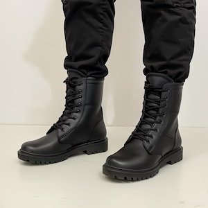 Puede incluir: Botas de combate de cuero negro con dise&ntilde;o de cordones. Las botas tienen una puntera redondeada y una suela gruesa con tacos. Las botas se llevan con pantalones negros.