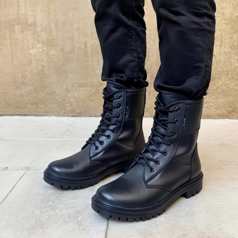 Puede incluir: Un par de botas de combate de cuero negro con dise&ntilde;o de cordones. Las botas tienen punta redonda, suela gruesa y altura media de la pantorrilla. Las botas se usan con pantalones negros.