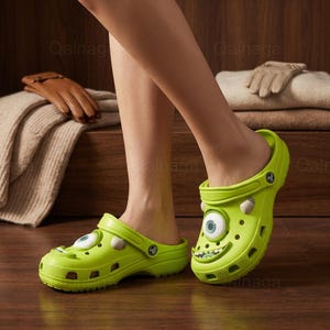 May include: Lime green monster-themed clogs with a cartoon eye and mouth design. The shoes have a strap and ventilation holes. The clogs are a fun and playful footwear option.
