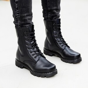 Puede incluir: Botas de combate de cuero negro con dise&ntilde;o de cordones. Las botas tienen una puntera redondeada y una suela gruesa. Las botas se llevan con vaqueros negros.