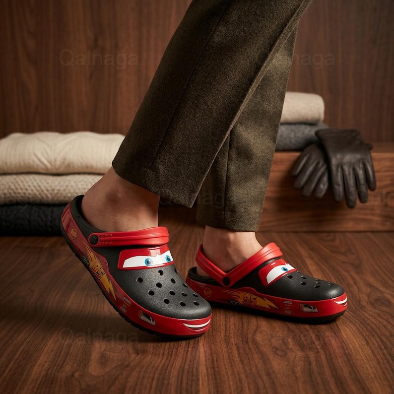 May include: Black and red Lightning McQueen Crocs clogs. The clogs feature a Lightning McQueen design with his face on the front and a red strap. The shoes are on a wooden floor.
