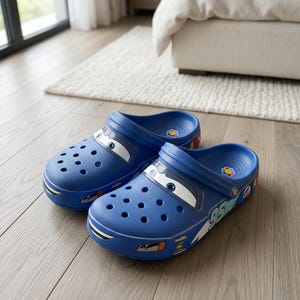 May include: A pair of blue slip-on clogs featuring a graphic print of a race car with white eyes on the toe box, the number 95 on the side, and a small colorful flower logo on the heel bed. The shoes have ventilation holes and a heel strap.
