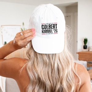 May include: A white baseball cap with the text "COLBERT KIMMEL '28" in black and red. The cap is being held up by a person with long blonde hair. The text is centered on the front of the cap.