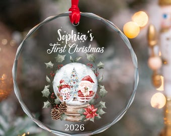 May include: A round, faceted glass Christmas ornament hanging from a red ribbon. It features the text 'Sophia's First Christmas' and '2026' above and below a graphic of a snow globe containing a snowman, Santa Claus, and a tree, framed by holly.