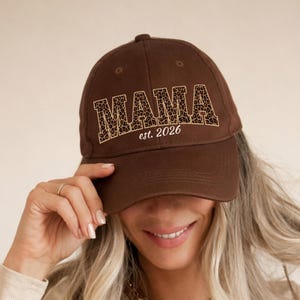 May include: A brown baseball cap with the word "MAMA" in a leopard print design and the text "est. 2026". The cap is a classic six-panel style with a curved brim. The hat is being worn by a person.
