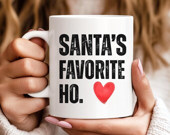 May include: White ceramic mug with the text "SANTA'S FAVORITE HO." in black, with a red heart. The mug has a curved handle and is held by a person.