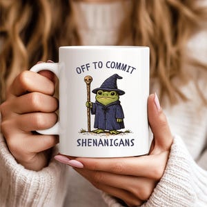 May include: White ceramic mug featuring a cartoon frog dressed as a wizard, holding a staff. The frog wears a blue robe and hat. Text on the mug reads "OFF TO COMMIT SHENANIGANS".