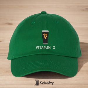 May include: Green baseball cap with the embroidered text "VITAMIN G" below a graphic of a pint of stout with a golden harp. The cap has a curved brim and a top button.